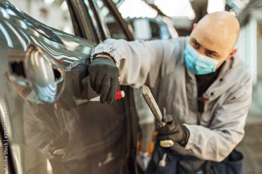 A car factory worker wearing a protective medical mask removes a metal defect with a hand tool. A car factory worker wearing a protective medical mask removes a metal defect with a hand tool.