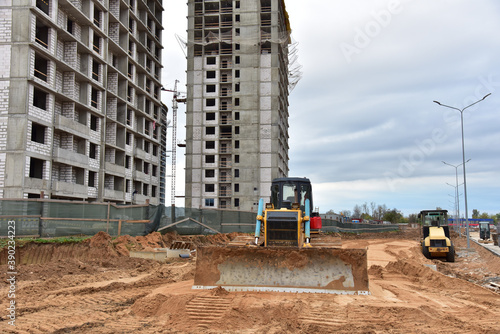 View of a large construction site where earthmoving equipment. Dozer and Soil compactor leveling ground for new road construction. Tower cranes are building tall residential buildings