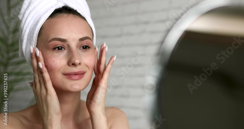 Beautiful woman with bare shoulders applying face cream after morning shower. Happy young lady with towel on head moisturising skin at home.