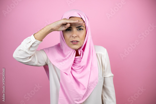 Young beautiful arab woman wearing islamic hijab over isolated pink background very happy and smiling looking far away with hand over head. Searching concept. © Irene