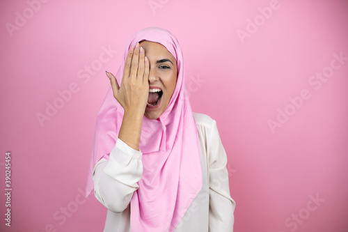 Young beautiful arab woman wearing islamic hijab over isolated pink background covering one eye with hand, confident smile on face and surprise emotion.