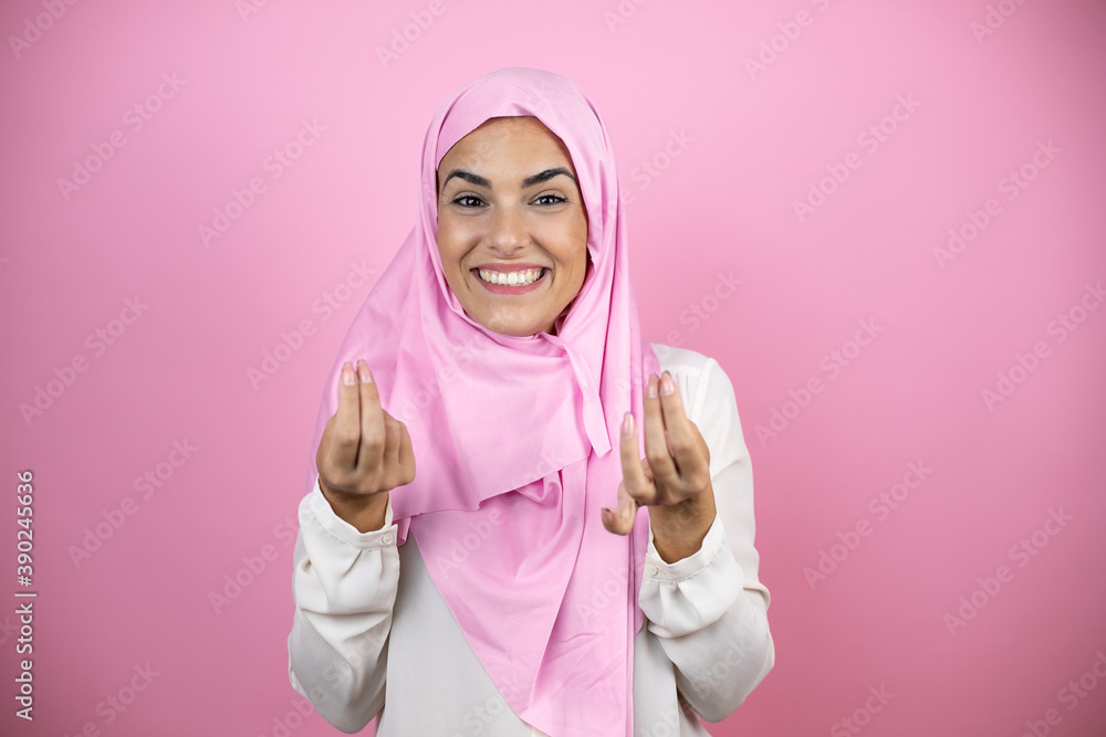 Young beautiful arab woman wearing islamic hijab over isolated pink background doing money gesture with hands, asking for salary payment, millionaire business Young beautiful arab woman wearing islamic hijab over isolated pink background doing money gesture with hands, asking for salary payment, millionaire business