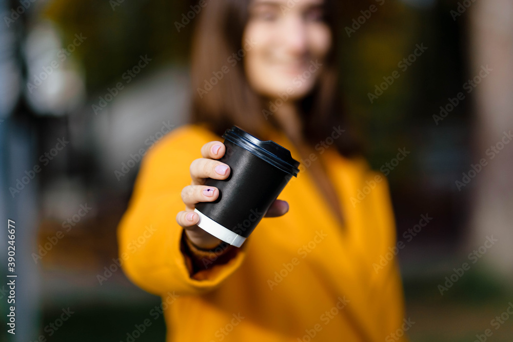 Female hand holding a cup of blank paper on a background of the park. Free space. Hands of a young girl in a yellow coat holding a paper cup of coffee. Girl hand hold coffee plastic cup. Coffee. Female hand holding a cup of blank paper on a background of the park. Free space. Hands of a young girl in a yellow coat holding a paper cup of coffee. Girl hand hold coffee plastic cup. Coffee.