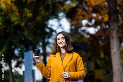 Female hand holding a cup of blank paper on a background of the park. Free space. Hands of a young girl in a yellow coat holding a paper cup of coffee. Girl hand hold coffee plastic cup. Coffee. © Alex