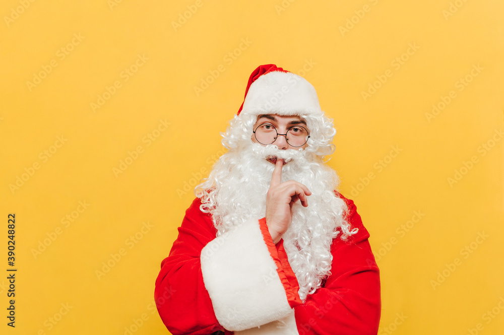 Serious Santa Claus shows a sign of silence and looks intently at the camera on a yellow background. Santa shows a finger to the lips gesture Shhh Serious Santa Claus shows a sign of silence and looks intently at the camera on a yellow background. Santa shows a finger to the lips gesture Shhh
