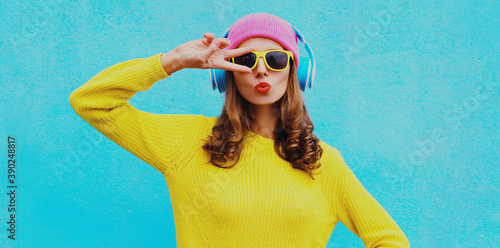 Portrait of modern young woman in wireless headphones listening to music wearing yellow knitted sweater and pink hat on blue background