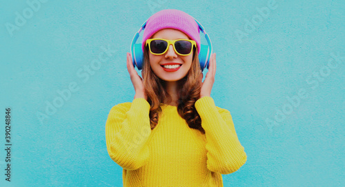 Portrait of happy smiling young woman in wireless headphones listening to music wearing yellow knitted sweater and pink hat on blue background