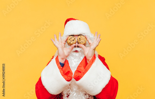 Funny santa with chocolate chip cookies in hands holding eyes near yellow background. Santa Claus and Christmas cookies, isolated. New Year