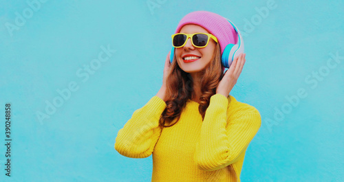 Portrait of happy smiling young woman in wireless headphones listening to music wearing yellow knitted sweater and pink hat on blue background