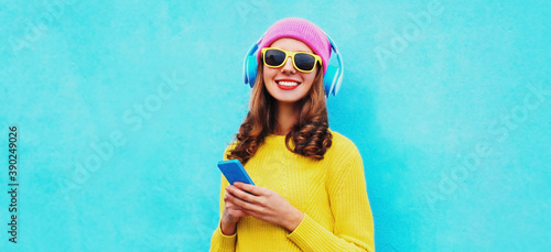 Portrait of modern young woman in wireless headphones listening to music with phone wearing yellow knitted sweater and pink hat on blue background