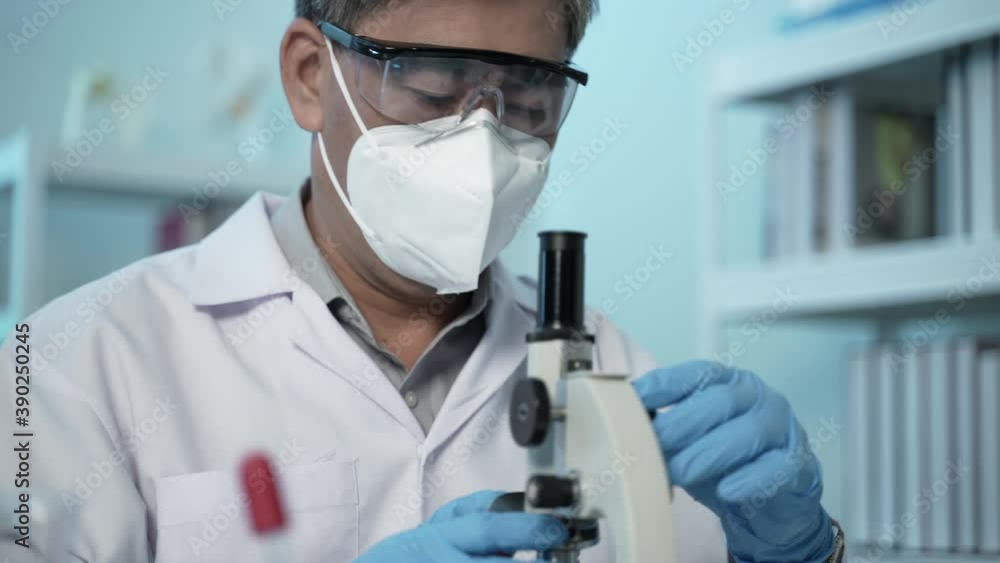 unrecognizable virologist in protective mask, suit and gloves looking through microscope while researching on Covid-19 treatment in laboratory with biohazard sign unrecognizable virologist in protective mask, suit and gloves looking through microscope while researching on Covid-19 treatment in laboratory with biohazard sign