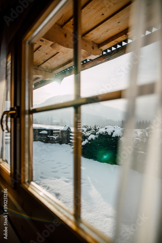 Wooden Cottage House Window. Beautiful snowy winter morning behind the chalet window. Beautiful mountain view. Sunshine and sparkling snow on fir trees in the cabin backyard. © AlexMaster