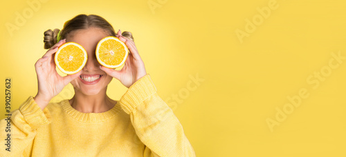 young blonde woman holding orange slice over her face, smiling on yellow background, wearing yellow sweater, with copy space 