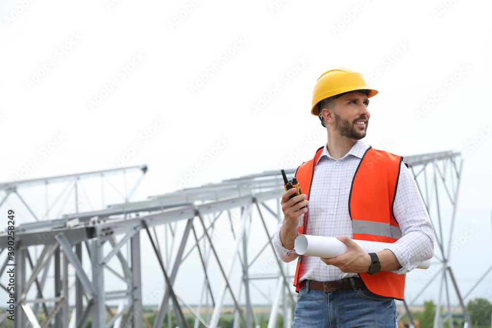 Professional engineer with plan and walkie talkie near high voltage tower construction outdoors. Installation of electrical substation Professional engineer with plan and walkie talkie near high voltage tower construction outdoors. Installation of electrical substation