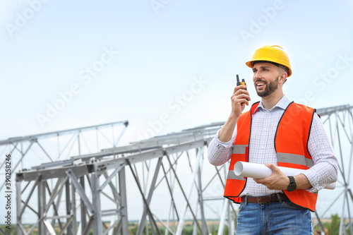 Professional engineer with plan and walkie talkie near high voltage tower construction outdoors. Installation of electrical substation