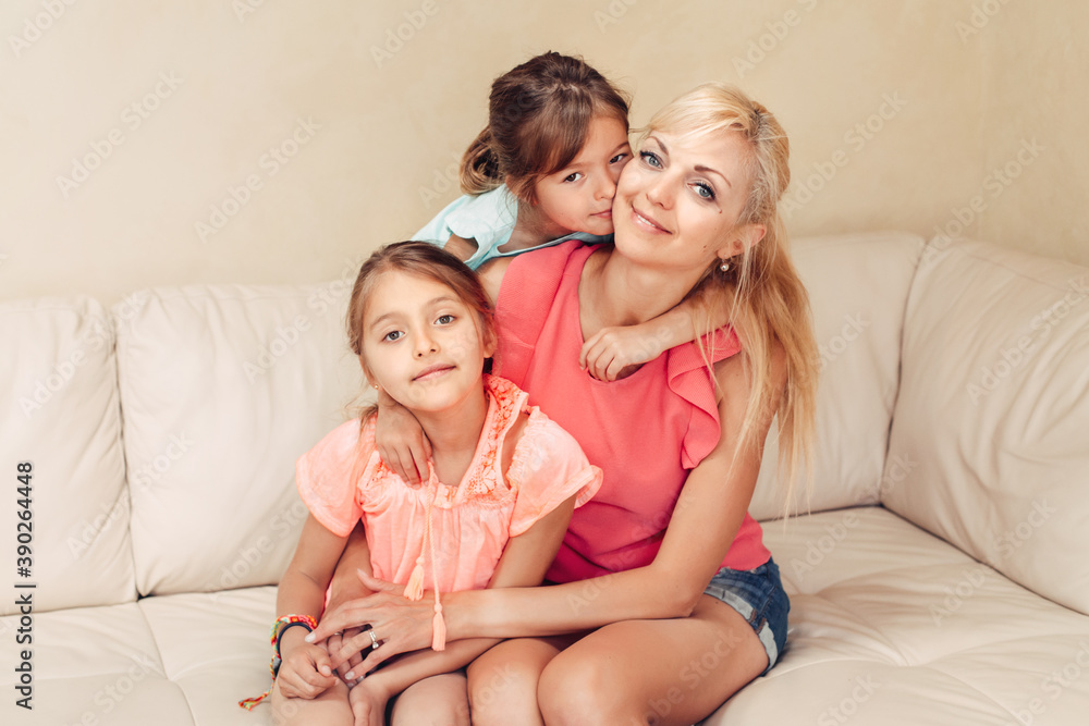 Mother and daughters girls hugging together on couch. Young Caucasian woman playing with children sisters at home. Family mom and two sibling spend time together. Mothers day holiday.