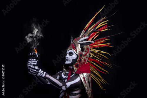 hombre latino , catrin de dios azteca, calavera prehispanica, con saumerio e incienso y fuego, portado penacho de plumas amarillas y rojas en fondo negro, dia de muertos, 1 de noviembre, calaca, photo