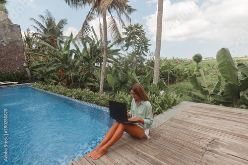 Woman working on her laptop computer sitting at poolside. Freelance work in tropical country © Yevhenii