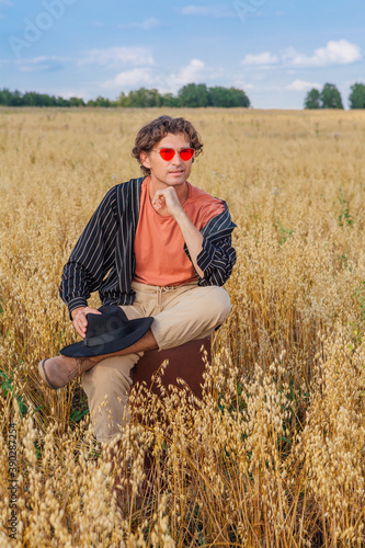 Tall handsome man with black hat and pink sunglasses sitting on a brown vintage leather suitcase at golden oat field.