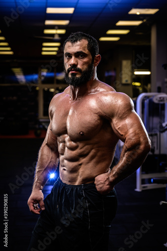 muscular strong young bearded caucasian male with powerful body in dark gym with blue and yellow lights