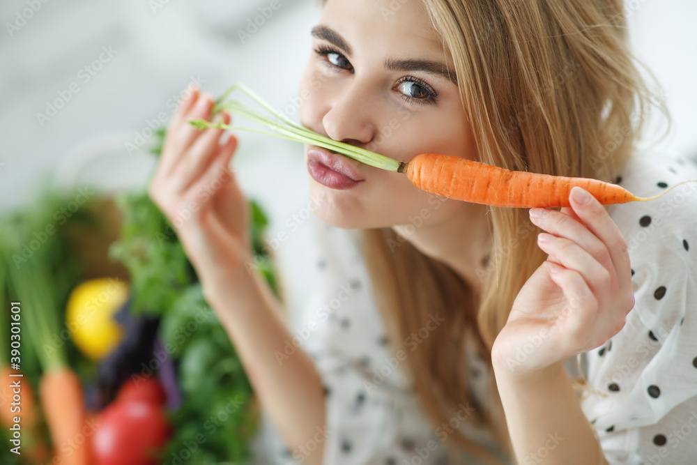 Young and happy woman with carrot at table with green fresh ingredients indoors. High quality photo. Young and happy woman with carrot at table with green fresh ingredients indoors. High quality photo.