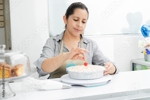 Hispanic woman in her patisserie decorating a cake- woman putting cherry on cake with whipped cream - enterprising woman