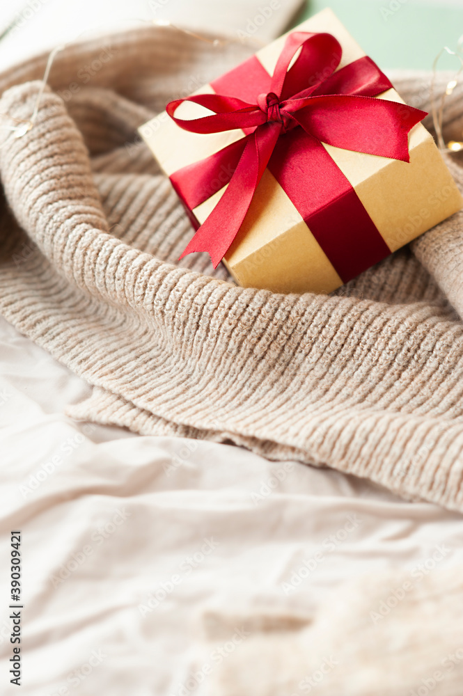 Red stylish square beige box with red festive satin ribbon on knitted beige cover with garlands. Cozy vertical holiday card with space for text Red stylish square beige box with red festive satin ribbon on knitted beige cover with garlands. Cozy vertical holiday card with space for text