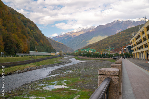 Beautiful alley on the river embankment on the background of ski resort and autumn mountains. Sochi, Krasnaya Polyana, Russia.