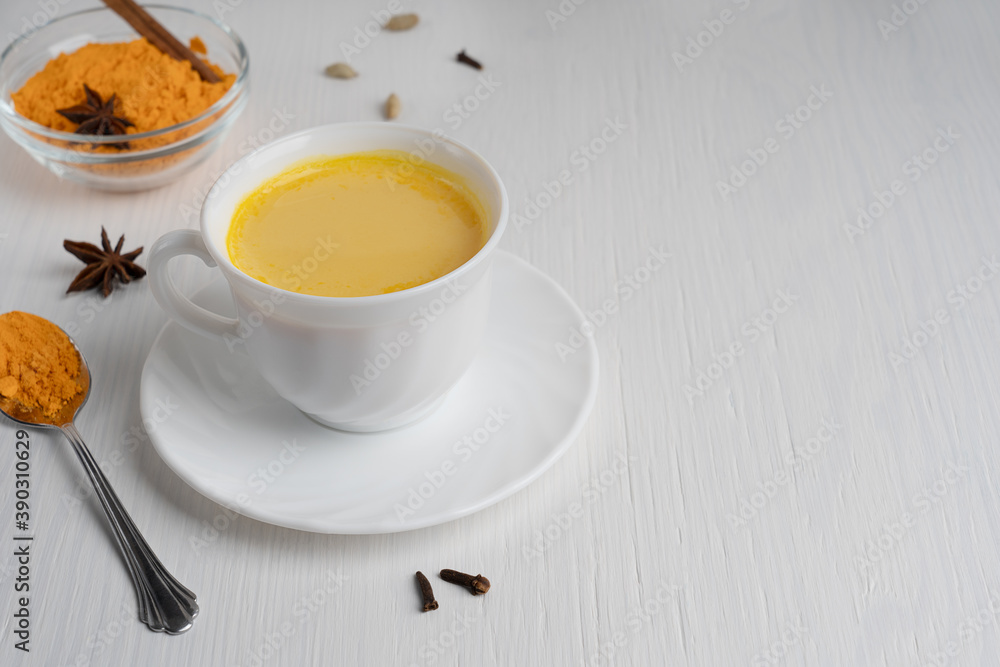Cup full of golden or turmeric milk which is healthy, healing drink having anti-inflammatory properties served on plate with spoon of curcuma on white wooden background. Image with copy space Cup full of golden or turmeric milk which is healthy, healing drink having anti-inflammatory properties served on plate with spoon of curcuma on white wooden background. Image with copy space