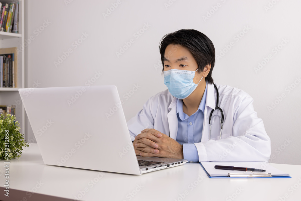 Young Asian Doctor Man in Lab Coat or Gown with Stethoscope Wear Face Mask Listening Patient Via Laptop Computer on Doctor Table in Office Young Asian Doctor Man in Lab Coat or Gown with Stethoscope Wear Face Mask Listening Patient Via Laptop Computer on Doctor Table in Office