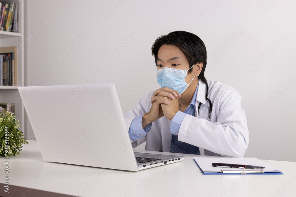 Young Asian Doctor Man in Lab Coat or Gown with Stethoscope Wear Face Mask and Thinking Front Laptop Computer on Doctor Table in Office Young Asian Doctor Man in Lab Coat or Gown with Stethoscope Wear Face Mask and Thinking Front Laptop Computer on Doctor Table in Office