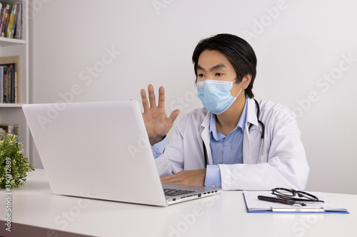 Young Asian Doctor Man in Lab Coat or Gown with Stethoscope Wear Face Mask Video Chat or Video Conference and Talking with Patient Via Laptop Computer on Doctor Table in Office