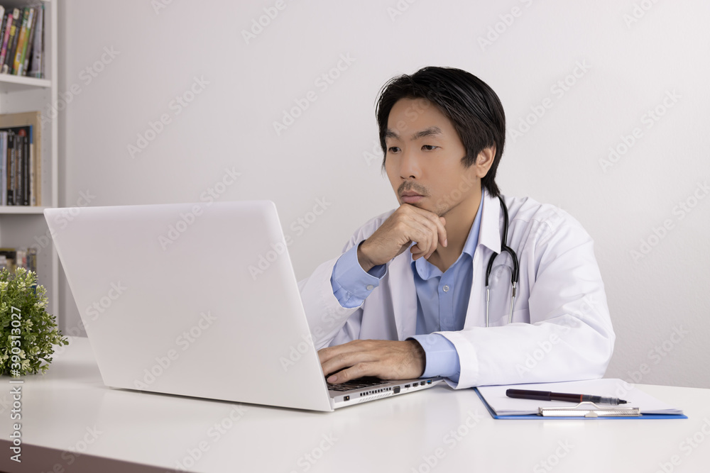 Serious Young Asian Doctor Man in Lab Coat or Gown with Stethoscope Touch Chin and Using Laptop Computer on Doctor Table in Office Serious Young Asian Doctor Man in Lab Coat or Gown with Stethoscope Touch Chin and Using Laptop Computer on Doctor Table in Office