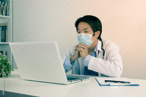 Young Asian Doctor Man in Lab Coat or Gown with Stethoscope Wear Face Mask and Thinking Front Laptop Computer on Doctor Table in Office in Vintage Tone