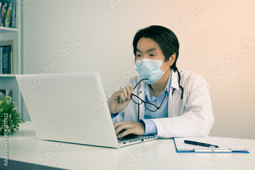 Young Asian Doctor Man in Lab Coat or Gown with Stethoscope Wear Face Mask and Holding Glasses in Serious Mood Using Laptop Computer on Doctor Table in Office in Vintage Tone