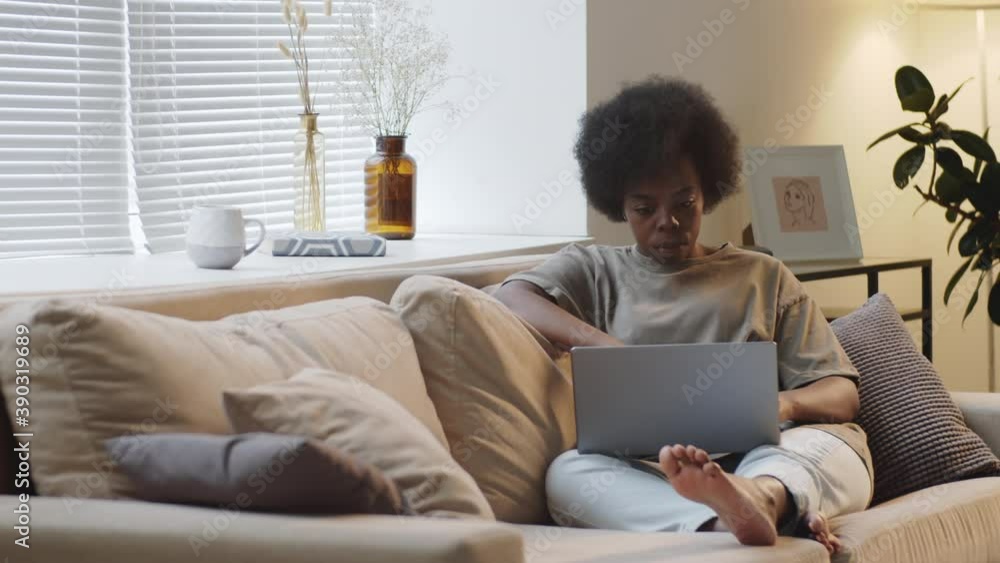 Young barefoot Afro-American woman sitting on sofa in the living room and using laptop while staying at home in isolation Young barefoot Afro-American woman sitting on sofa in the living room and using laptop while staying at home in isolation
