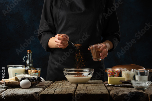 The chef in black apron pours cocoa into glass bowl for making dough. Backstage of cooking waffle on rustic wooden table with ingredients on dark blue background. Frozen motion. Traditional recipe.