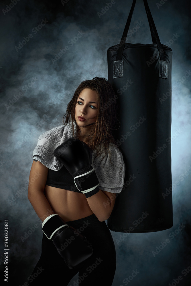 confident sporty woman in black boxing gloves near punching bag on dark studio background with smoke