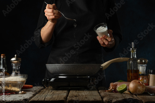 Professional chef pours some soda into frying pan for cooking traditional indian curry. Backstage of preparing on dark blue background. Concept of organizing the cooking process. Frozen motion.