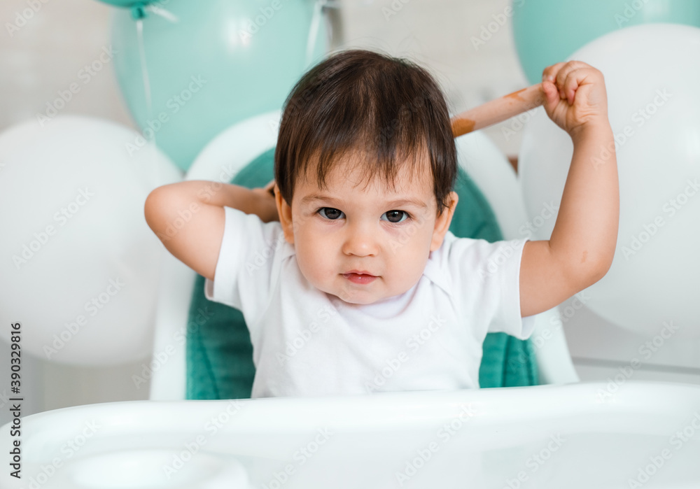 Little baby boy sitting in blue high chair at home on white kitchen and playing with wooden big spoon on background with balloons. Little baby boy sitting in blue high chair at home on white kitchen and playing with wooden big spoon on background with balloons.