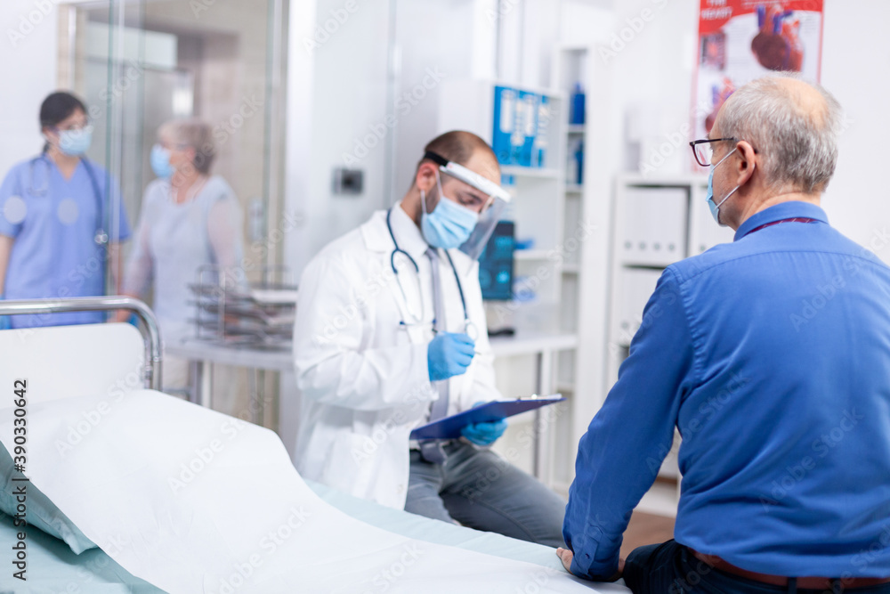Doctor taking notes on clipboard wearing protective mask against coronavirus outbreak during consultation of senior woman in hospital examination room. Practitioner physician appointment. Doctor taking notes on clipboard wearing protective mask against coronavirus outbreak during consultation of senior woman in hospital examination room. Practitioner physician appointment.