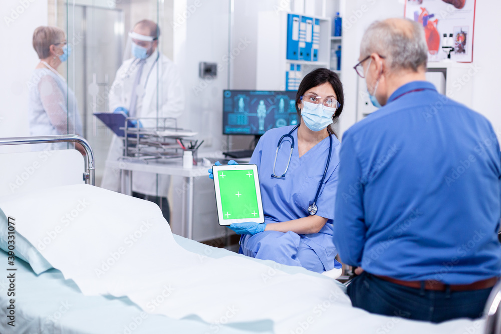 Nurse with face mask for protection against covid 19 holding tablet with green mockup while consulting senior man. Medical examination for infections, disease and diagnosis. Nurse with face mask for protection against covid 19 holding tablet with green mockup while consulting senior man. Medical examination for infections, disease and diagnosis.