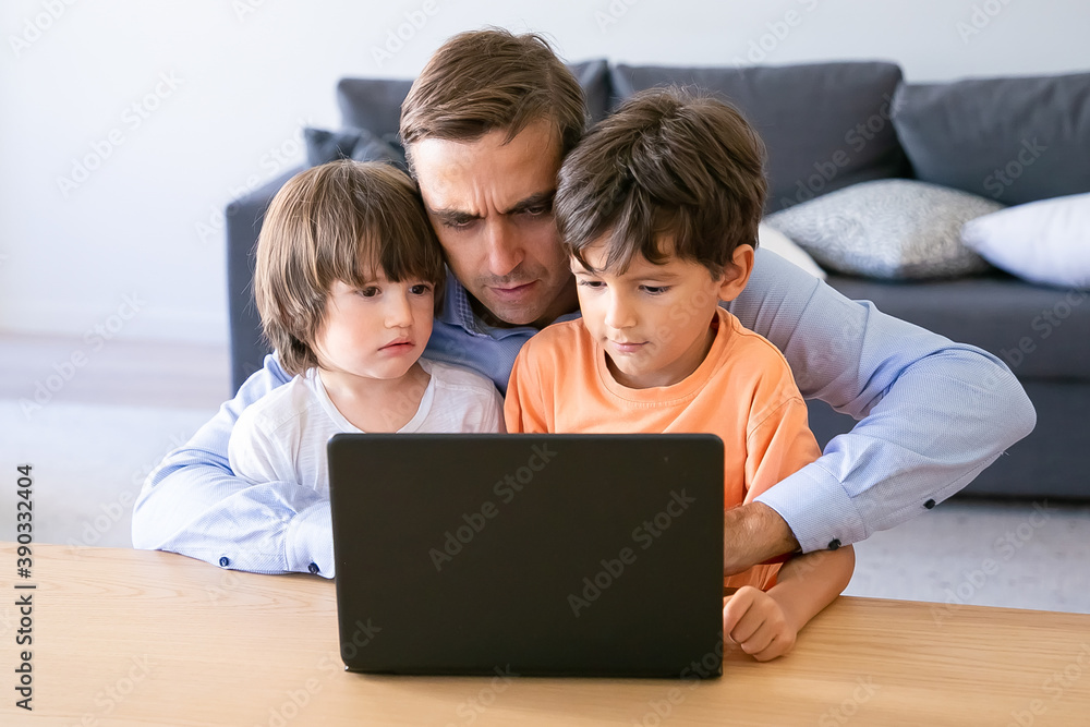 Serious dad working on laptop and embracing sons. Concentrated Caucasian father using laptop at home. Two cute boys sitting on his knees. Fatherhood, childhood and digital technology concept
