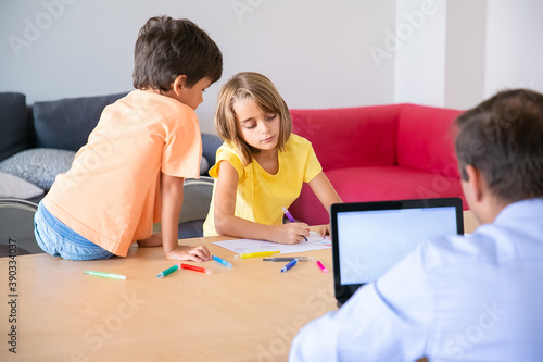 Caucasian dad working on laptop and cute kids painting doodles at table. Concentrated blonde girl drawing with marker and brother looking at her. Childhood, creativity and weekend concept