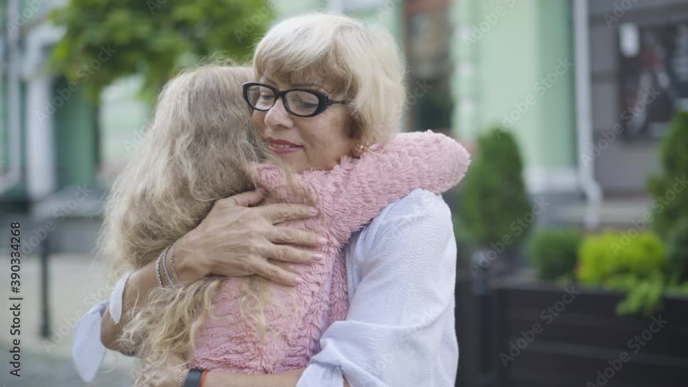 Camera moving around hugging grandmother and granddaughter. Happy smiling senior Caucasian woman in eyeglasses embracing little curly-haired girl on sunny urban city street. Camera moving around hugging grandmother and granddaughter. Happy smiling senior Caucasian woman in eyeglasses embracing little curly-haired girl on sunny urban city street.