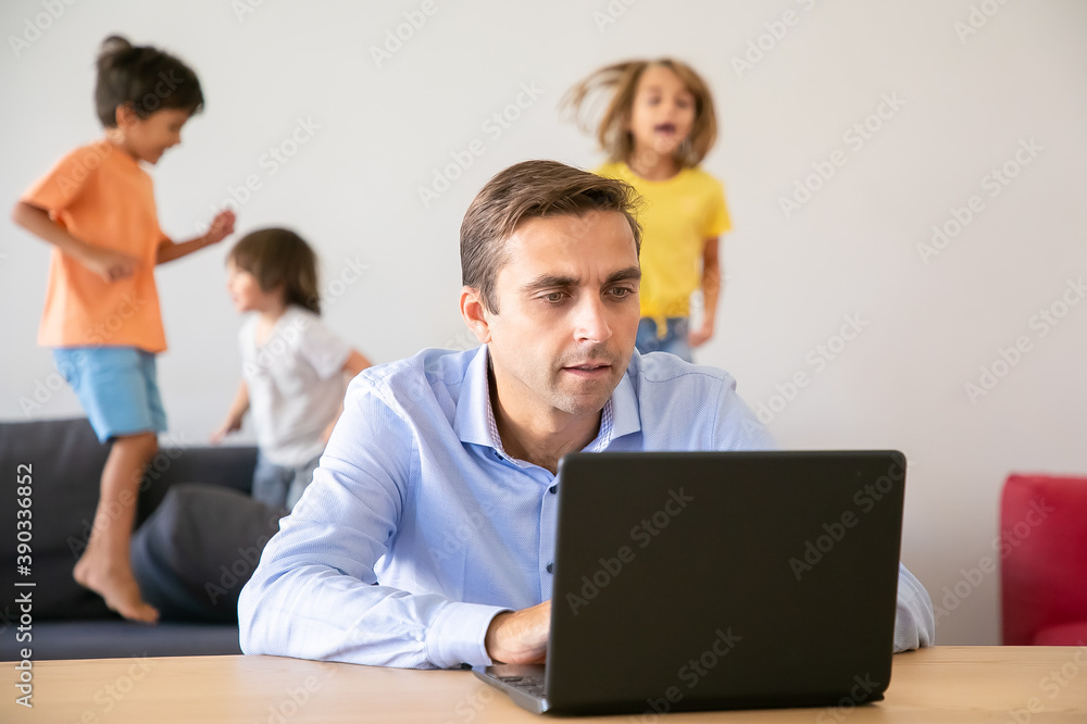 Serious Caucasian dad working via laptop and kids jumping on background. Concentrated father using computer and kids playing on sofa. Selective focus. Childhood and digital technology concept Serious Caucasian dad working via laptop and kids jumping on background. Concentrated father using computer and kids playing on sofa. Selective focus. Childhood and digital technology concept