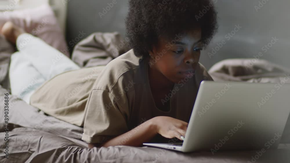 Young black woman lying on bed and using laptop while staying at home during lockdown Young black woman lying on bed and using laptop while staying at home during lockdown
