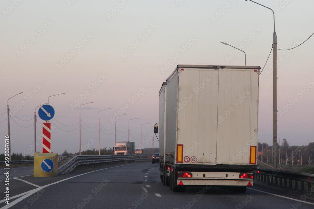 European white van truck with semi trailer drive on two lane suburban empty asphalted highway , back view at summer evening on blue pink sky background, transportation logistics road traffic