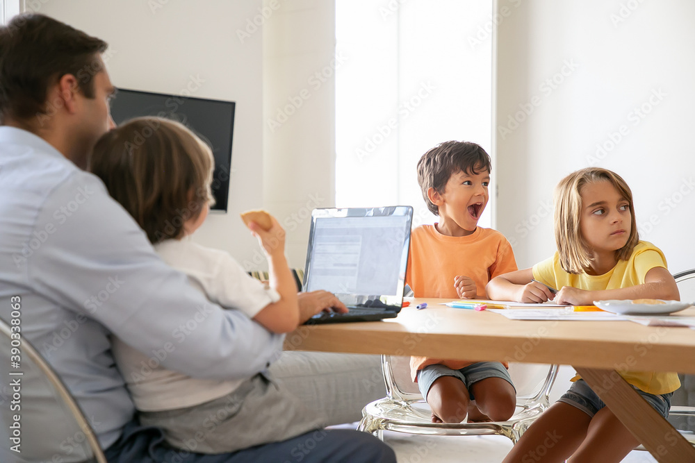 Happy kids drawing doodles when dad working on laptop and holding son on knees. Exited children painting on paper. Caucasian family sitting at table. Childhood, creativity and weekend concept Happy kids drawing doodles when dad working on laptop and holding son on knees. Exited children painting on paper. Caucasian family sitting at table. Childhood, creativity and weekend concept