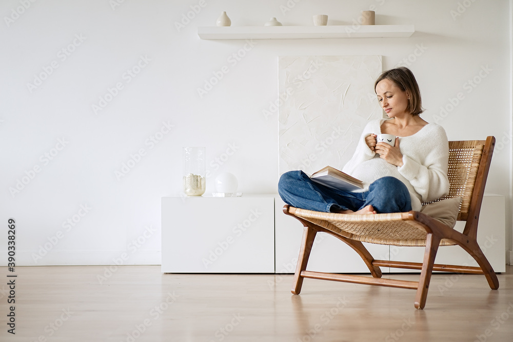 Beautiful pregnant woman sitting in an armchair by the window, reading a book and drinking tea Beautiful pregnant woman sitting in an armchair by the window, reading a book and drinking tea
